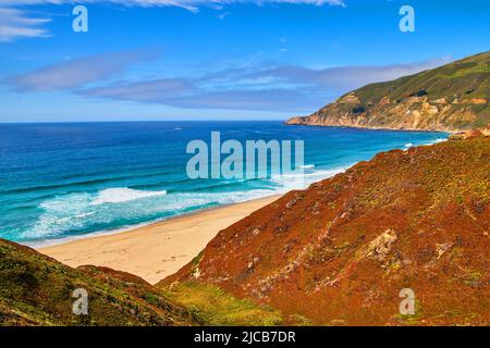 Friedliche Wellen treffen auf den Sandstrand neben den Hügeln mit frühlingshaften roten und grünen Pflanzen Stockfoto