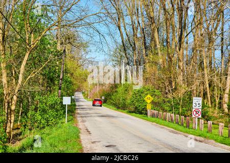 Rotes Auto, das im späten Frühling im mittleren Westen die Seitenstraße entlang fährt und Blätter aufbläst Stockfoto