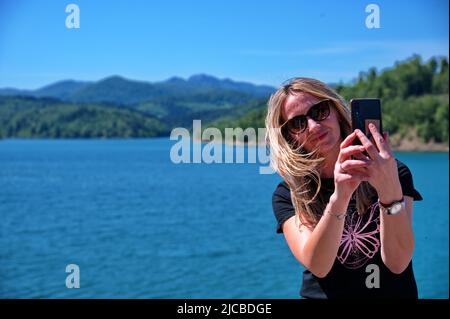 Reife Frau, die am Seeufer sitzt und mit dem Handy telefoniert Stockfoto