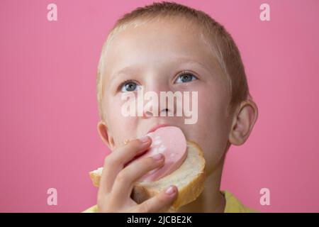 Junge isst ein Sandwich mit gekochter Wurst und sieht interessiert aus Stockfoto