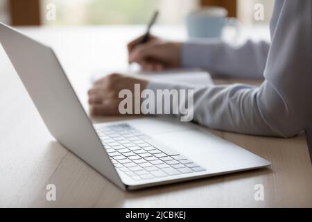 Unbekannte Frau sitzt am Schreibtisch mit einem Laptop, der auf Papier jottet Stockfoto