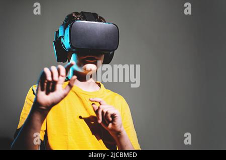 Netter Junge mit 3D Brillen-Headset, der Inhalte im Metaverse-Cyberspace beobachtet - isoliert auf schattiertem Hintergrund mit Kopierraum Stockfoto