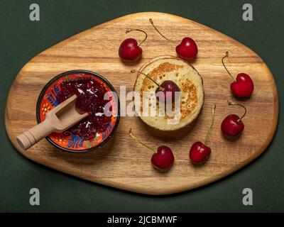 Kirschmarmelade, rote Kirschen und Pfannkuchen auf einem Holzschneidebrett Stockfoto