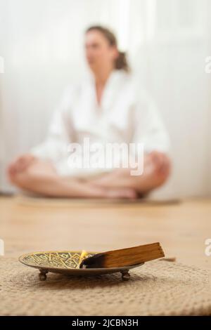 Brennend mit gelber Flamme palo santo Holzstab auf einem Metalltablett und Jute Seil Platzmatte, mit einer verschwommenen Frau meditieren in Lotus Pose verschwommen in der Stockfoto