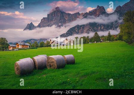 Majestätisches Bergdorf und Heuballen auf den blühenden grünen Wiesen. Atemberaubende hohe Nebelberge bei Sonnenuntergang, Dolomiten, San Vito di Cadore, Italien, Euro Stockfoto