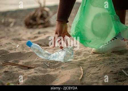 Nicht erkennbare afrikanische Frau, die sich hinlehnt, um Müll vom Sand am Strand in einer grünen Plastiktüte zu sammeln. Ökologie, Umweltverschmutzung. Stockfoto