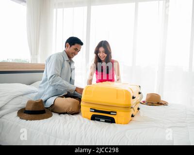 Paar Reisende, die sich auf die Reise vorbereiten. Asiatischer Mann im Denim-Hemd und junge schöne sitzen zusammen und packen Tasche, gelbe Koffer auf dem Bett im Schlafzimmer. Stockfoto
