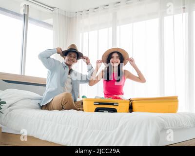 Paar Reisende bereit zu reisen. Junge asiatische Frau und Mann im Denim-Hemd tragen Strandhüte, während sie mit gelben Koffern im Schlafzimmer packen und sie betrachten Stockfoto