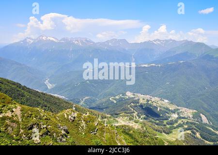 Berglandschaft mit schneebedeckten Bergen, blauem Himmel und Wolken. Berghang bedeckt mit grünem Gras und Tannen. Stockfoto