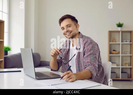 Fröhlicher Student, der mit einem Laptop und einem Notebook am Schreibtisch sitzt, eine Brille hält und lächelt Stockfoto