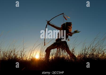 Ein junges indianisches Mädchen der Ureinwohner Amerikas wird bei Sonnenuntergang im hohen Gras gesehen. Sie wird gesehen, wie sie mit Silhouetten ihren Pfeil und Bogen schießt, während die Sonne hinter ihr untergeht. Stockfoto