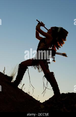 Ein junges indianisches Mädchen spielt bei Sonnenuntergang Flöte. Das Mädchen ist im Schatten, als die Sonne hinter ihr untergeht. Stockfoto