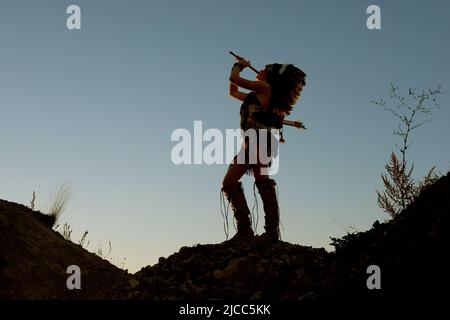 Ein junges indianisches Mädchen spielt bei Sonnenuntergang Flöte. Das Mädchen ist im Schatten, als die Sonne hinter ihr untergeht. Stockfoto