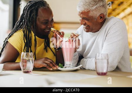 Glückliches reifes Paar lächelt einander an, während sie einen leckeren Milchshake teilen. Fröhliches Seniorenpaar, das sich in einem Restaurant gut Vergnügen hat. Carefree Matte Stockfoto