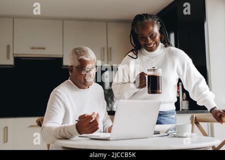 Eine reife Frau lächelt glücklich, während sie im Büro ihres Mannes eine Kanne Kaffee hält. Unterstützende ältere Frau bringt ihrem Mann ein heißes Getränk woh Stockfoto