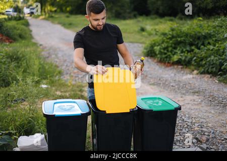 Mann wirft Plastikflasche in den Recyclingbehälter auf dem Hinterhof. Verschiedene Farben auf Kunststoffbehältern. Grün, blau und gelb Stockfoto