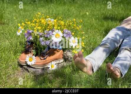 Braune Schuhe mit Blumenstrauß in ihnen, ruhendes Kind liegt auf Gras in der Nähe. Blume Überraschung Geschenk für Mama. Aktiver Lebensstil, Spaziergänge, Liebe zur na Stockfoto