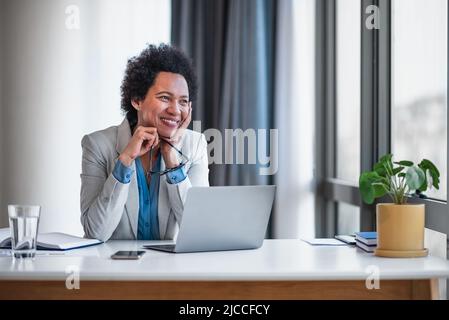 Eine besonnene Geschäftsfrau mit Lächeln blickt weg, während sie am Laptop arbeitet. Weibliche professionelle in formellen sitzt am Schreibtisch. Sie erwägt in offi Stockfoto