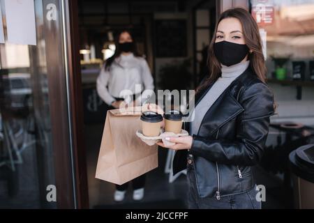 Attraktive Kundin in Gesichtsmaske steht im Freien beim Café und nimmt Essen und Kaffee mit. Hintergrund des Kassierers in Maske wartet auf nächste Stockfoto
