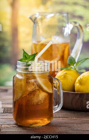 Glas Eistee mit Zitronenscheiben, Minze und Eis mit Zitronen in einer Schüssel und Krug im Hintergrund auf einem rustikalen Holztisch Stockfoto