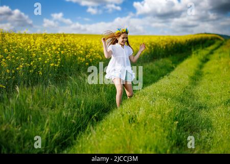 Schönes, energiegeladenes, verträumtes Teenager-Mädchen in einem weißen Kleid und einem leuchtend ukrainischen Kranz, das durch gelb blühende Rapsfelder und grüne Äcker läuft Stockfoto