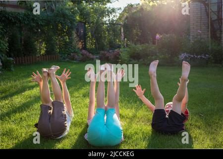 Drei barfuss-Kinder liegen mit hochgestreckten Beinen und hochgestreckten Armen auf dem Gras. Fröhlicher Spaziergang im Park an einem sonnigen Sommertag. Sommerstimmung, glückliche Kindheit Stockfoto