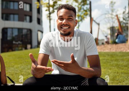 Positiver schwarzer Mann, der Videoanruf macht, spricht und gestikuliert zur Kamera, im Stadtgebiet im Freien sitzt und lächelt Stockfoto