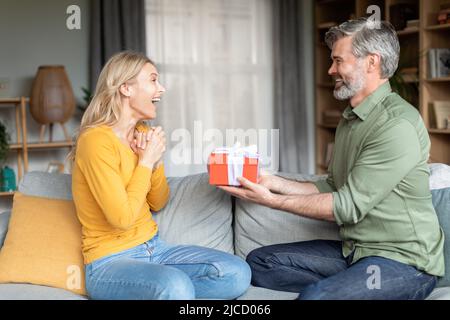 Liebevoller Mann mittleren Alters, der seiner aufgeregten Frau zu Hause ein Geschenk macht Stockfoto