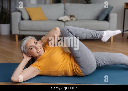 Aktive ältere Frau, die zu Hause auf einer Yogamatte ABS-Übungen macht und die Kamera anlächelt und im Wohnzimmer trainiert Stockfoto