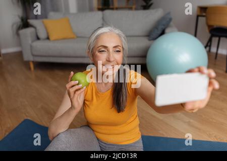 Inländischer Sport und gesunde Ernährung. Ältere Frau, die Selfie mit Apfel macht und sich nach dem Heimtraining auf der Fitnessmatte ausruht Stockfoto