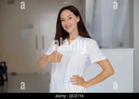 Porträt einer lächelnden Krankenschwester, die in der Nähe der Rezeption in einer medizinischen Klinik steht und den Daumen nach oben zeigt Stockfoto