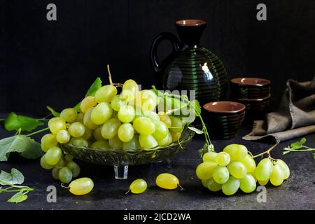 Frische reife Trauben mit Blättern in einer Obstschale und Keramikglas und Gläsern auf schwarzem Hintergrund Stockfoto