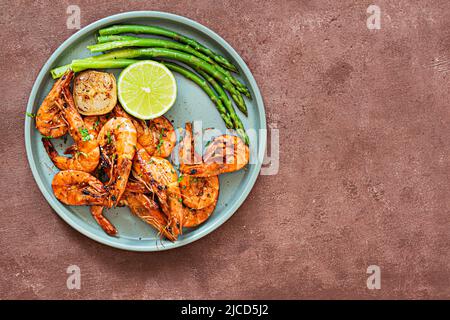 Gegrillte Garnelen (Tigergarnelen) in Gewürzen mit Spargel und einer Scheibe Limette (Zitrone) auf einem Teller auf braunem Hintergrund. Stockfoto