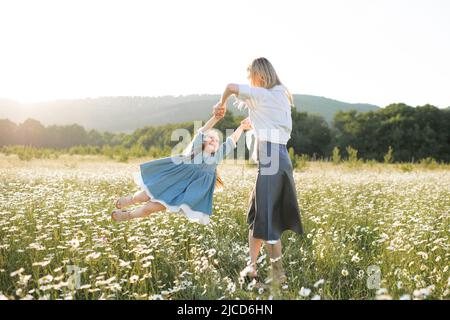 Nette schöne junge Frau hält die Hände und spielen mit kleinen Mädchen gehen in Kamillenfeld mit blühenden Blumen über Natur Hintergrund. Familie Stockfoto