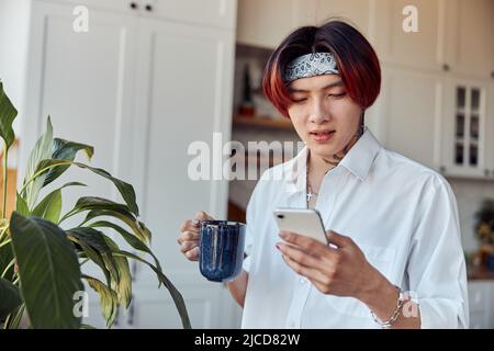 Stilvoller asiatischer Kerl mit Telefon, während er eine Tasse Kaffee in seiner Wohnung hält Stockfoto