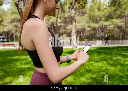 Schöne Brünette Frau trägt Sport-BH auf Stadtpark stehen, im Freien Hände halten Telefon berühren Finger Mockup weißen leeren Display, mobile App Stockfoto