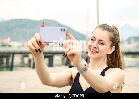 Nette kaukasische Frau in sportlicher Kleidung im Stadtpark, im Freien, die mit ihrem Smartphone ein horizontales Foto macht. Selektiver Fokus auf ihr Gesicht. Stockfoto