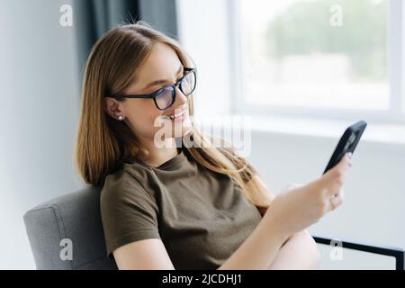 Schöne Nachricht von ihm. Schöne junge lächelnde Frau, die ihr Smartphone anschaut, während sie in einem großen bequemen Stuhl zu Hause sitzt Stockfoto