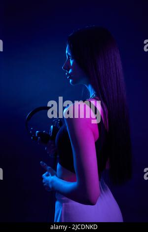 Anmutiges langhaariges Mädchen, das Kopfhörer hält und gerade im dunklen Studio steht. Blick von der Schulter des Brünetten Weibchens in schwarzer Spitze mit Ohrhörern, hervorgehoben durch dunkles magentafarbenes Licht. Konzept der Kunst Stockfoto