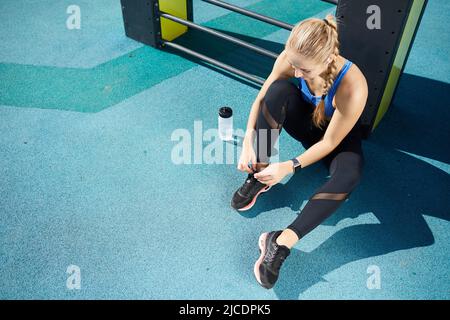 Schöne junge Frau mit blondem Zopf sitzt auf dem Boden im Freien und binden Sportschuh Spitze Stockfoto