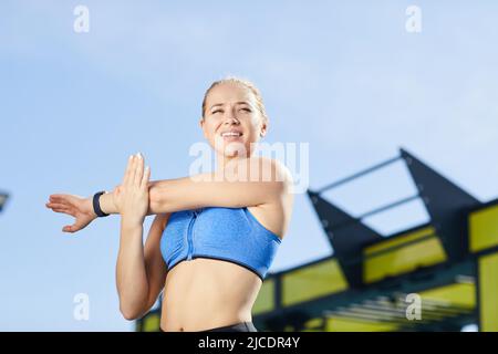 Jolly energische junge Frau im Sport-BH, die auf dem Außengelände steht und nach dem Training Bizeps streckt Stockfoto