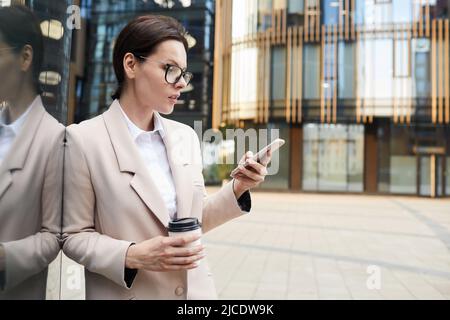 Nachdenkliche, moderne Geschäftsfrau mittleren Alters in Brillen, die an der Gebäudewand gelehnt sind und Smartphone verwenden, während sie mit dem Geschäftspartner kommuniziert Stockfoto