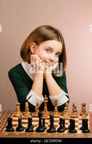 Portrait Cute Schulmädchen mit kurzen Haaren sitzt am Schachbrett. Junge Schachspielerin Kindermädchen tragen grünes Kleid, das vor dem Schachspiel posiert. Stockfoto