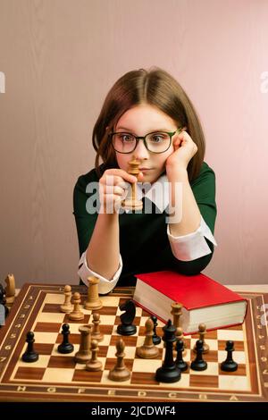 Portrait Lustige spielerische junge Schachspielerin mit kurzem Haarschnitt in grünem Kleid. Schulmädchen in Brillen sitzen am Schachbrett und geschlossen Red Pape Stockfoto