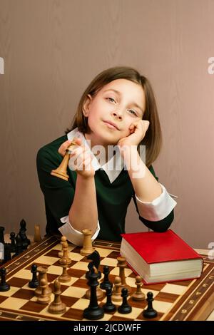 Portrait stolze selbstbewusste junge Schachspielerin mit kurzem Haarschnitt in grünem Kleid. Schulmädchen sitzt am Schachbrett und schließt rotes Papierbuch, Stockfoto