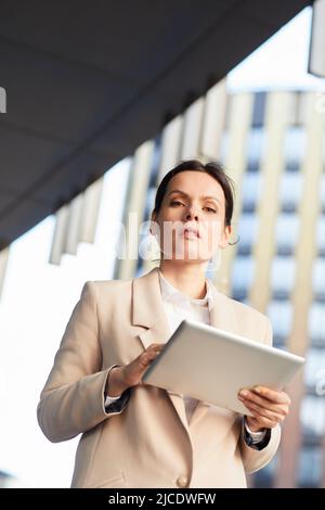 Unten sehen Sie eine ernsthafte, strenge Geschäftsanalytikerin mit braunem Haar, die ein digitales Tablet für die Arbeit verwendet, während sie über die Straße geht Stockfoto