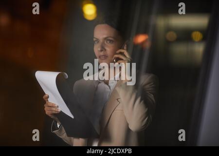 Nachdenkliche Dame mittleren Alters in der Jacke hält die Zwischenablage mit Papieren und diskutiert den Vertrag mit dem Lieferanten am Telefon Stockfoto