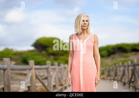 Schöne reife Frau, die einen Holzweg in der Nähe des Strandes entlang geht, in einem schönen orangefarbenen Kleid. Stockfoto