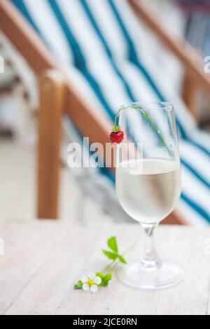 Ein Glas Weißwein mit Erdbeere auf dem Hintergrund eines Liegestuhls am Strand. Sommerzeit. Sommerliche Stimmung. Stockfoto