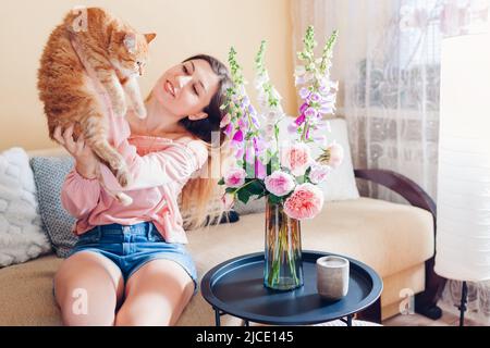Spielen mit Katze zu Hause durch frischen Strauß. Junge Frau sitzt und entspannt auf der Couch im Wohnzimmer und hält Haustier. Sich wohlfühlen mit Kätzchen Stockfoto